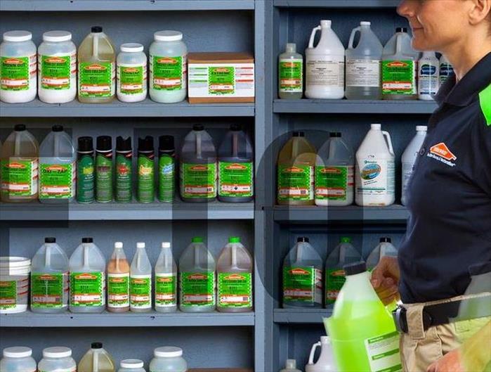woman in a hat with a ponytail carries a bottle of cleaner in front of shelves filled with cleaning products