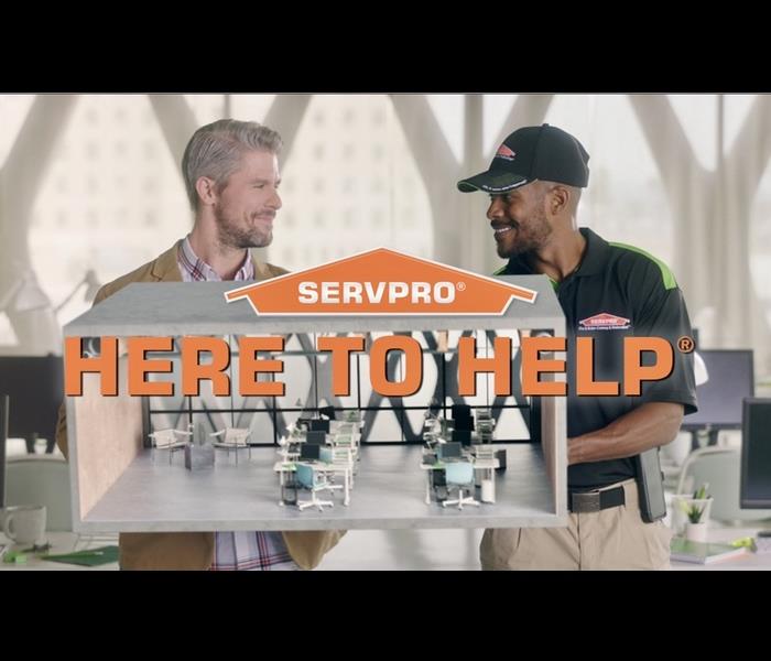 man in SERVPRO uniform and a man in a suit jacket stand behind a model of an office, large orange letters say 'Here to Help'